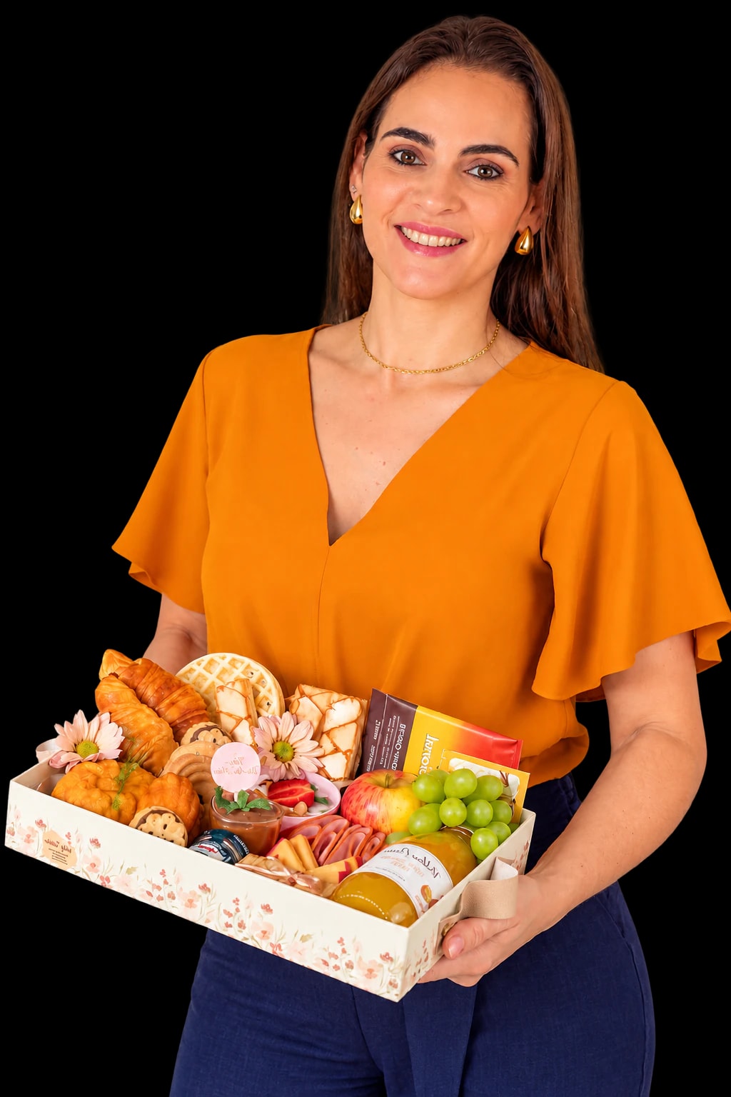 Gaby Tenório sorrindo, de blusa laranja, segurando uma cesta de café da manhã com croissants, frutas e flores